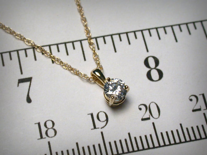 May include: A gold-colored necklace with a small, round, clear gemstone pendant. The chain is delicate and the pendant is set in a four-prong setting. The necklace is displayed against a white background with a ruler.