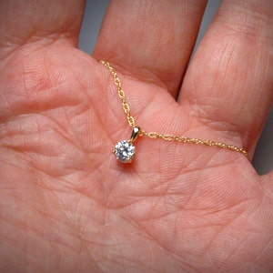 May include: A delicate gold necklace featuring a small, round, clear gemstone pendant. The pendant is set in a gold setting and hangs from a thin, gold chain. The necklace is displayed against a neutral background, highlighting the jewelry's simple elegance.
