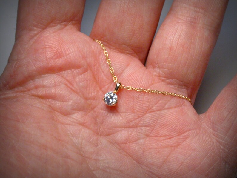 May include: A delicate gold necklace featuring a small, round, clear gemstone pendant. The pendant is set in a gold setting and hangs from a thin, gold chain. The necklace is displayed against a neutral background, highlighting the jewelry's simple elegance.