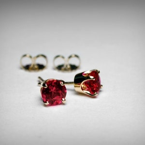 May include: A pair of gold stud earrings with red gemstones. The earrings have a simple design and are perfect for everyday wear.