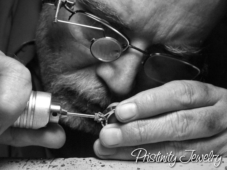 May include: Black and white image of a jeweler working on a ring. The jeweler is wearing glasses and using a small tool to refine the ring's details. The text "Pristinity Jewelry" is visible at the bottom of the image.