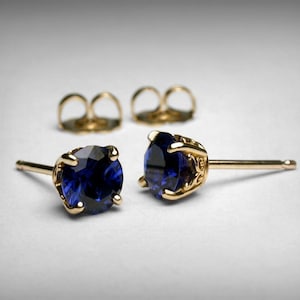 May include: A pair of gold stud earrings with deep blue gemstones. The earrings have a classic design with a four-prong setting. Two gold earring backs are also visible.
