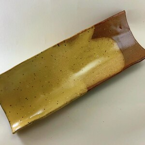 May include: A long, rectangular ceramic serving dish with a speckled, light yellow glaze on one side and a brown glaze on the other side.