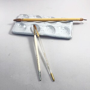 Puede incluir: Una paleta de pintura de cer&aacute;mica blanca con nueve pozos, un pincel de madera y dos pinceles finos. La paleta tiene una textura moteada.