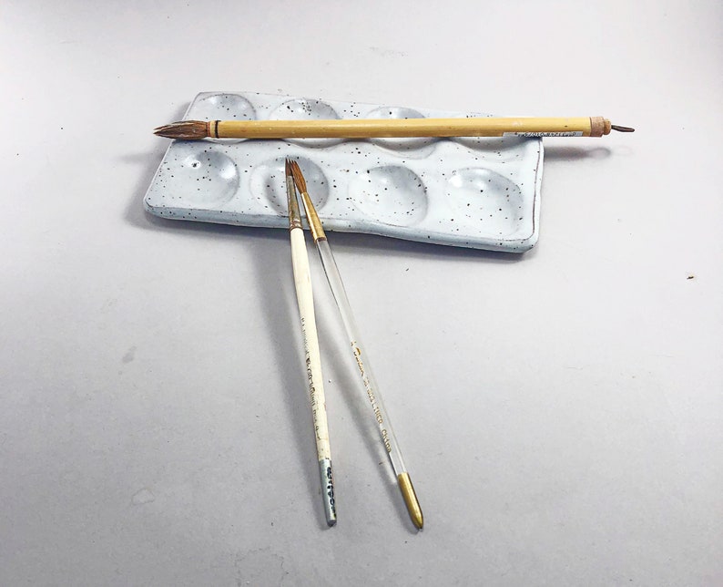 Puede incluir: Una paleta de pintura de cer&aacute;mica blanca con nueve pozos, un pincel de madera y dos pinceles finos. La paleta tiene una textura moteada.