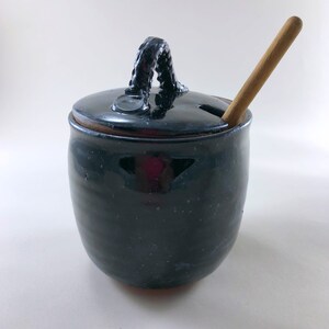 May include: Black ceramic honey pot with a lid and a wooden honey dipper. The pot has a textured handle and a small hole in the lid for the dipper.