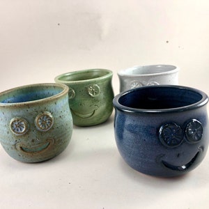 May include: Four ceramic mugs with different colored glazes and smiling faces. The mugs are all different colors, including green, blue, and white.