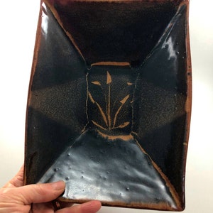 May include: A square ceramic dish with a dark brown glaze and a subtle metallic sheen. The dish has a raised design in the center, featuring a stylized plant motif in a lighter brown color.