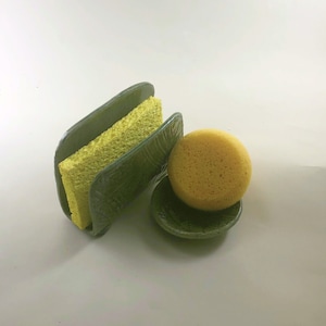 May include: A green ceramic dish with a yellow sponge inside and a yellow sponge sitting on top of a smaller green ceramic dish.