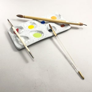 Puede incluir: Una paleta de pintura de cer&aacute;mica blanca con nueve pocillos, llenos de diferentes colores de pintura. Tres pinceles est&aacute;n sobre la paleta, dos son de madera y uno es transparente con una punta dorada.