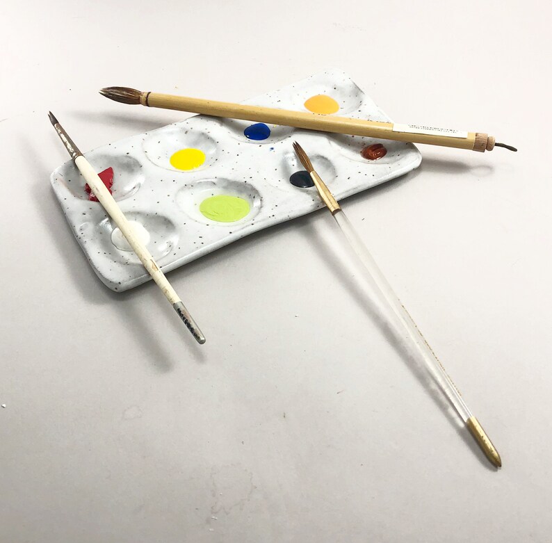Puede incluir: Una paleta de pintura de cer&aacute;mica blanca con nueve pocillos, llenos de diferentes colores de pintura. Tres pinceles est&aacute;n sobre la paleta, dos son de madera y uno es transparente con una punta dorada.