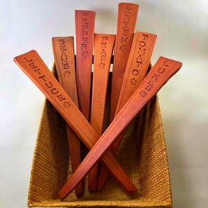 May include: A set of eight wooden plant markers with different vegetable names engraved on them. The markers are arranged in a woven basket.
