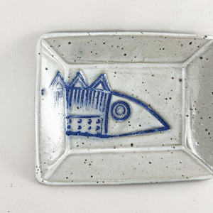 May include: A small, white ceramic dish with a blue fish design. The fish has a stylized, geometric design with a blue and white pattern.