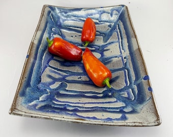 May include: A square ceramic serving dish with a blue and gray glaze, holding three red and orange peppers. The dish has a textured surface and a brown rim. The peppers have green stems.