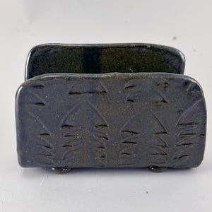May include: A dark charcoal gray ceramic napkin holder with a glossy finish and a speckled texture. The exterior features a repeating carved pattern of stylized leaf or feather shapes. It stands on four small, rounded ceramic feet.