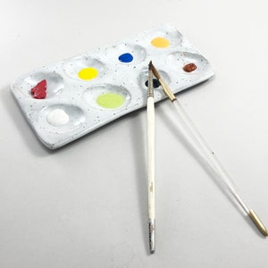 Puede incluir: Una paleta de pintura de cer&aacute;mica blanca con nueve pozos, llenos de pintura roja, amarilla, azul, verde y marr&oacute;n. Dos pinceles est&aacute;n sobre la paleta, uno con mango blanco y otro con mango transparente.