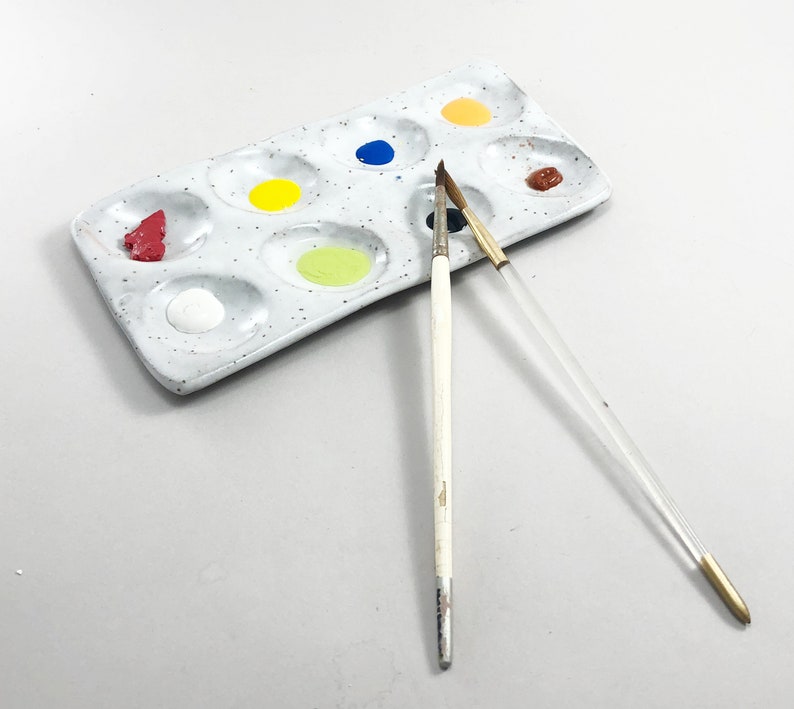 Puede incluir: Una paleta de pintura de cer&aacute;mica blanca con nueve pozos, llenos de pintura roja, amarilla, azul, verde y marr&oacute;n. Dos pinceles est&aacute;n sobre la paleta, uno con mango blanco y otro con mango transparente.
