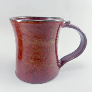 May include: A handmade ceramic mug with a reddish-brown glaze. The mug has a wide base and a slightly flared rim. The handle is attached to the side of the mug.