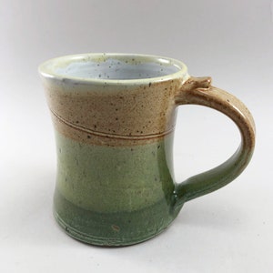 May include: A handmade ceramic mug with a light brown, green, and white glaze. The mug has a wide base and a rounded handle.