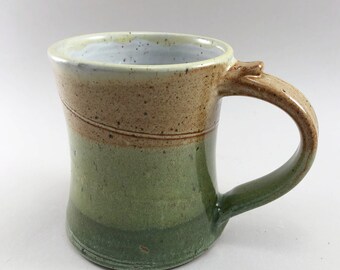 May include: A handmade ceramic mug with a light brown, green, and white glaze. The mug has a wide base and a rounded handle.