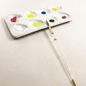 Puede incluir: Una paleta de pintura de cer&aacute;mica blanca con 10 pocillos, llenos de varios colores de pintura, incluyendo rojo, amarillo, azul, negro y marr&oacute;n. Un pincel delgado con mango de madera est&aacute; acostado junto a la paleta.