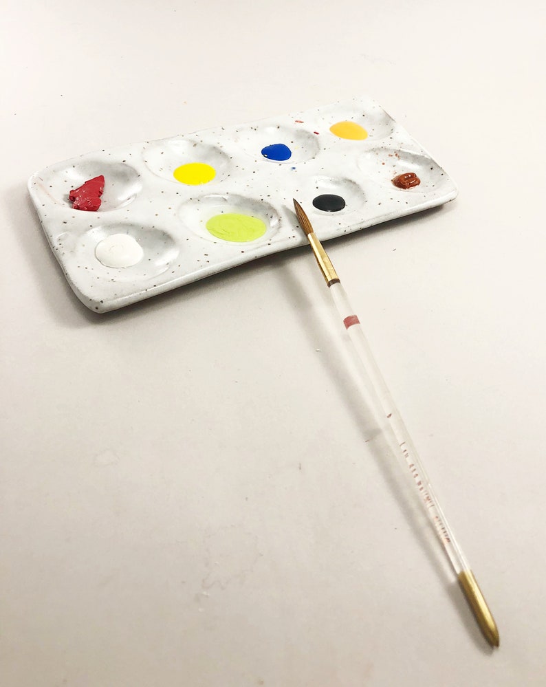 Puede incluir: Una paleta de pintura de cer&aacute;mica blanca con 10 pocillos, llenos de varios colores de pintura, incluyendo rojo, amarillo, azul, negro y marr&oacute;n. Un pincel delgado con mango de madera est&aacute; acostado junto a la paleta.