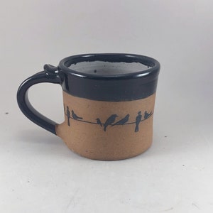 Puede incluir: Una taza de cerámica marrón y negra con un borde negro y un asa negra. La taza tiene un diseño de pájaros negros en un alambre en la parte delantera.