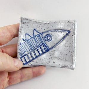 May include: A small, square ceramic dish with a hand-drawn blue fish design. The fish has a detailed eye, fins, and body markings. The dish is speckled gray with a slightly uneven surface, suggesting a handmade quality. The dish is held in a hand.