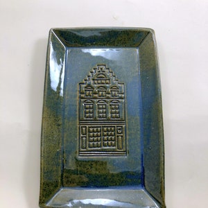 May include: A rectangular ceramic serving tray with a raised design of a building with windows and a roof. The tray is glazed in shades of blue and green.