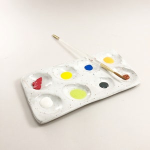 Puede incluir: Una paleta de pintura de cer&aacute;mica blanca con nueve pozos, llenos de pintura roja, amarilla, verde, azul, blanca y marr&oacute;n. Un pincel descansa sobre la paleta.