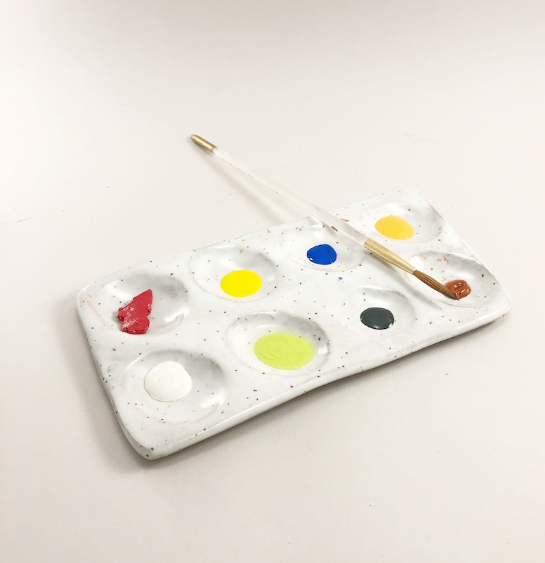 Puede incluir: Una paleta de pintura de cer&aacute;mica blanca con nueve pozos, llenos de pintura roja, amarilla, verde, azul, blanca y marr&oacute;n. Un pincel descansa sobre la paleta.