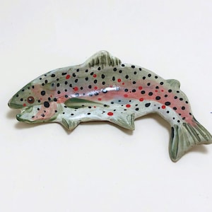 May include: A ceramic trout dish with a green, white, and red speckled design. The dish is shaped like a trout and has a raised tail and fins.