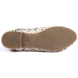 May include: A pair of women's flats with a cowhide pattern. The shoes have a white base with black spots and a tan sole. The sole is made of a textured material. The flats are designed for casual wear.