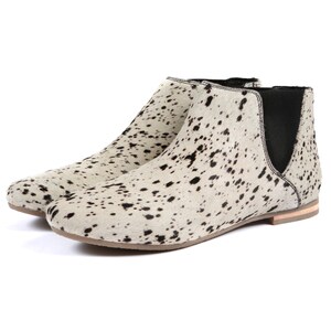 May include: A pair of ankle boots with a cowhide pattern. The boots have a white base with black spots, a rounded toe, and elastic side panels. The soles are a light brown color.