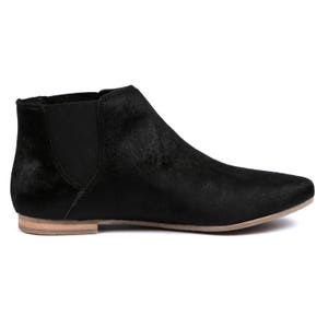May include: A black ankle boot with a pointed toe and a low heel. The boot features a textured, fur-like material and elastic side panels for easy wear. The sole is a light tan color.