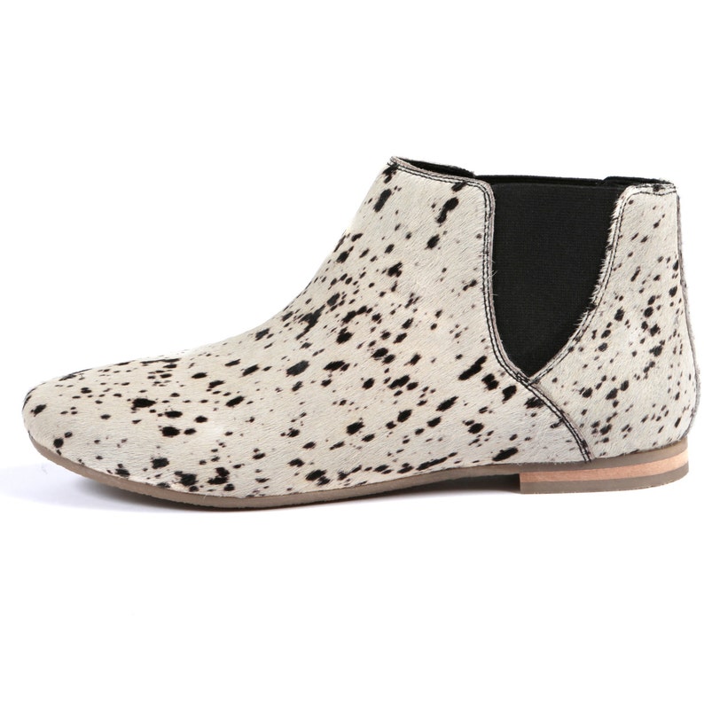 May include: A side view of a white and black spotted ankle boot. The boot has a rounded toe, a black elastic side panel, and a light brown sole. The boot's pattern resembles cowhide.