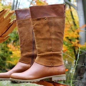 May include: A pair of knee-high brown leather boots with a suede panel and a zip closure. The boots are set against a blurred background of autumn foliage and a weathered wooden branch.