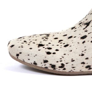 May include: Close-up of a shoe with a cowhide pattern. The shoe features a white background with black spots and a brown sole. The design resembles a Dalmatian dog's coat.