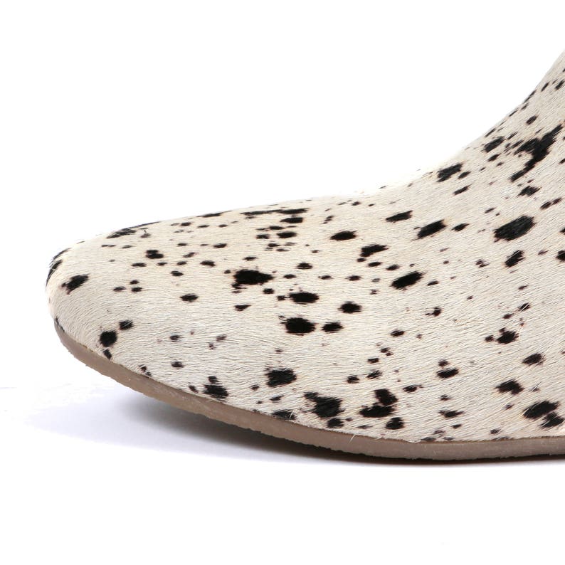 May include: Close-up of a shoe with a cowhide pattern. The shoe features a white background with black spots and a brown sole. The design resembles a Dalmatian dog's coat.