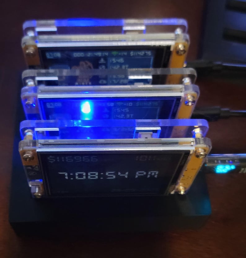 K&ouml;nnte beinhalten: Ein Stapel von vier kleinen elektronischen Ger&auml;ten mit transparenten Acrylgeh&auml;usen und blauen Lichtern. Jedes Ger&auml;t zeigt Daten auf einem kleinen Bildschirm an, wobei der untere Bildschirm "$116966" und "7:08:54 PM" anzeigt.