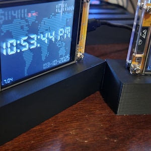 May include: Two digital clocks displaying the time 22:53:44 and 22:53:48, with the date 03-10-2025. The clocks have a black base and a clear frame with gold accents. The display shows the time and date, with a world map background.