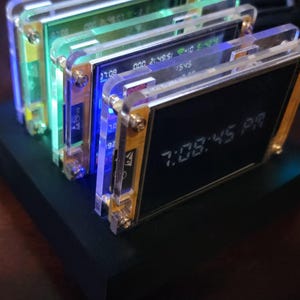 K&ouml;nnte beinhalten: Ein Stapel elektronischer Ger&auml;te mit transparenten Acrylrahmen und goldfarbener Hardware. Die Bildschirme zeigen Zeit und andere Daten an, wobei die Uhrzeit 19:08:45 auf einem angezeigt wird. Die Ger&auml;te stehen auf einer schwarzen Basis mit blauer und gr&uuml;ner Innenbeleuchtung.
