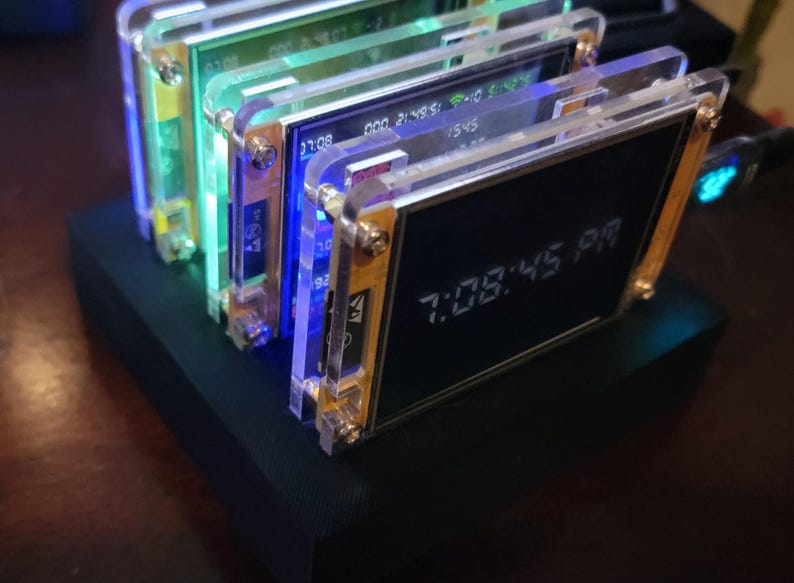 K&ouml;nnte beinhalten: Ein Stapel elektronischer Ger&auml;te mit transparenten Acrylrahmen und goldfarbener Hardware. Die Bildschirme zeigen Zeit und andere Daten an, wobei die Uhrzeit 19:08:45 auf einem angezeigt wird. Die Ger&auml;te stehen auf einer schwarzen Basis mit blauer und gr&uuml;ner Innenbeleuchtung.