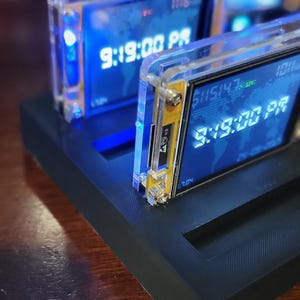 May include: Two digital clocks displaying the time 9:19:00 PM. The clocks have a blue display with white numerals and are encased in clear acrylic. They sit on a black base.