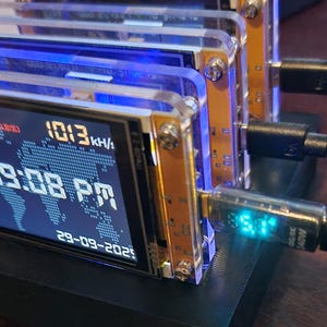 K&ouml;nnte beinhalten: Ein Stapel kleiner elektronischer Ger&auml;te mit transparenten Acrylgeh&auml;usen und goldfarbenen Leiterplatten. Jedes Ger&auml;t zeigt Zeit, Datum und andere Daten auf einem blauen Bildschirm an. USB-Kabel sind eingesteckt. Die Ger&auml;te sind auf einer schwarzen Basis montiert.