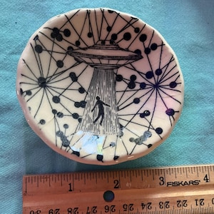 May include: Small, off-white ceramic dish with a black and white UFO design. The illustration depicts a flying saucer beaming up a person. The dish is approximately 3 inches in diameter.