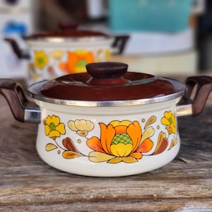 May include: A white enameled metal cooking pot with a dark brown lid and side handles. The exterior features a decorative floral pattern in shades of orange, yellow, and brown. A second, similar pot is visible in the blurred background.