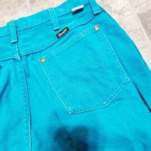May include: Close-up of turquoise Wrangler jeans. The jeans feature a classic five-pocket design with a branded patch and brass-colored rivets. The back pocket has a stitched 'W' design.