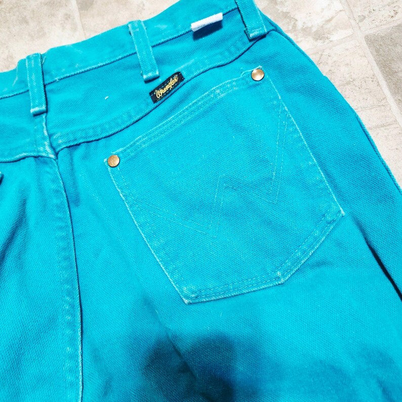 May include: Close-up of turquoise Wrangler jeans. The jeans feature a classic five-pocket design with a branded patch and brass-colored rivets. The back pocket has a stitched 'W' design.