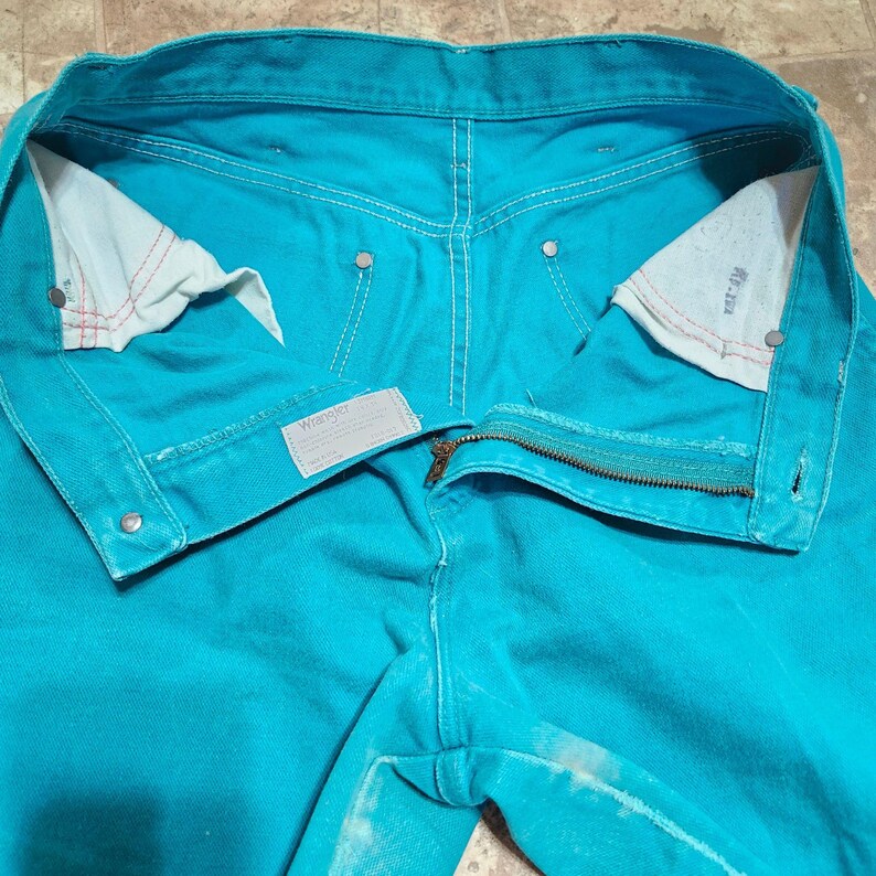 May include: Close-up of turquoise Wrangler jeans, unbuttoned and unzipped, showcasing the interior pockets and stitching. The jeans have a classic five-pocket design with a zipper fly and metal rivets. The Wrangler label is visible.